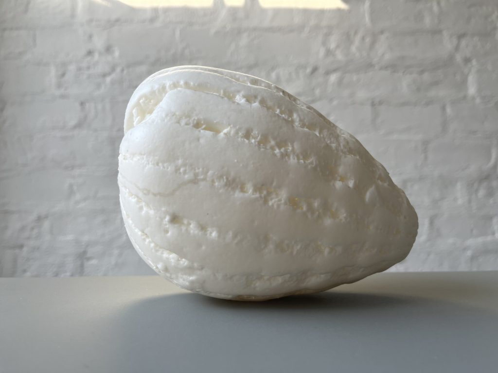 A white, textured, 3D-printed resin object sits on a gray surface against a white brick wall.