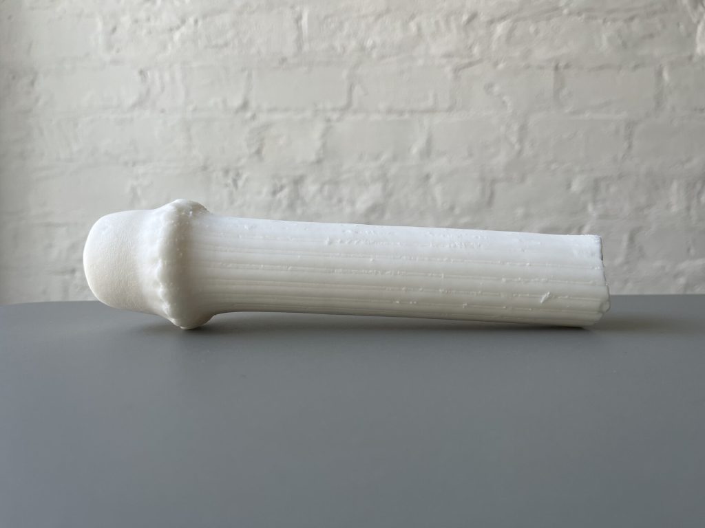 A white, 3D-printed resin object with a ribbed shaft and bulbous head sits on a gray surface against a white brick wall.