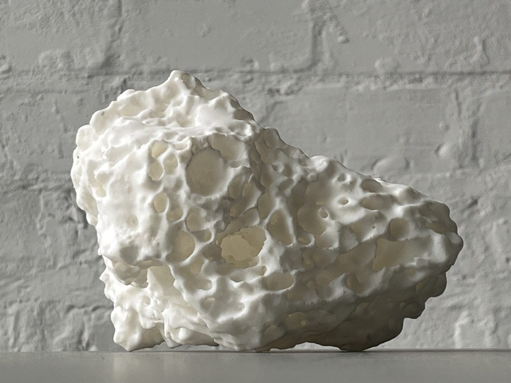A white, porous 3D-printed resin object sits on a surface against a textured wall.