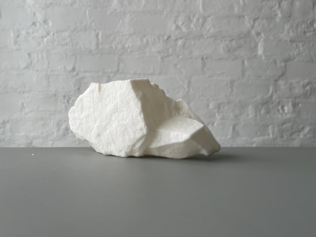 A white, textured 3D-printed resin object sits on a gray surface against a white brick wall.