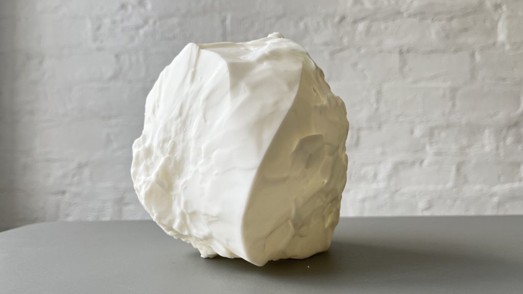 A white 3D-printed resin sculpture with textured, irregular surfaces sits on a gray surface against a white brick wall.