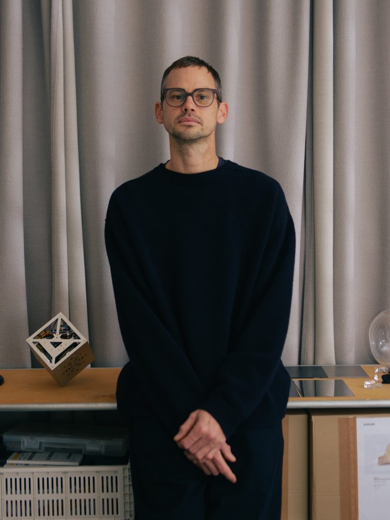A man in a dark sweater stands before a curtain with a wooden cube on a table to his left.