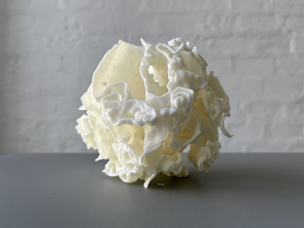 A white 3D-printed resin object with porous, coral-like structure sits on a gray surface against a white brick wall.