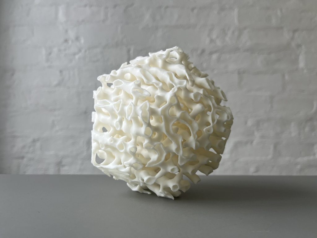 A white 3D-printed resin porouhedron with porous, bone-like structure sits on a gray surface against a brick wall.