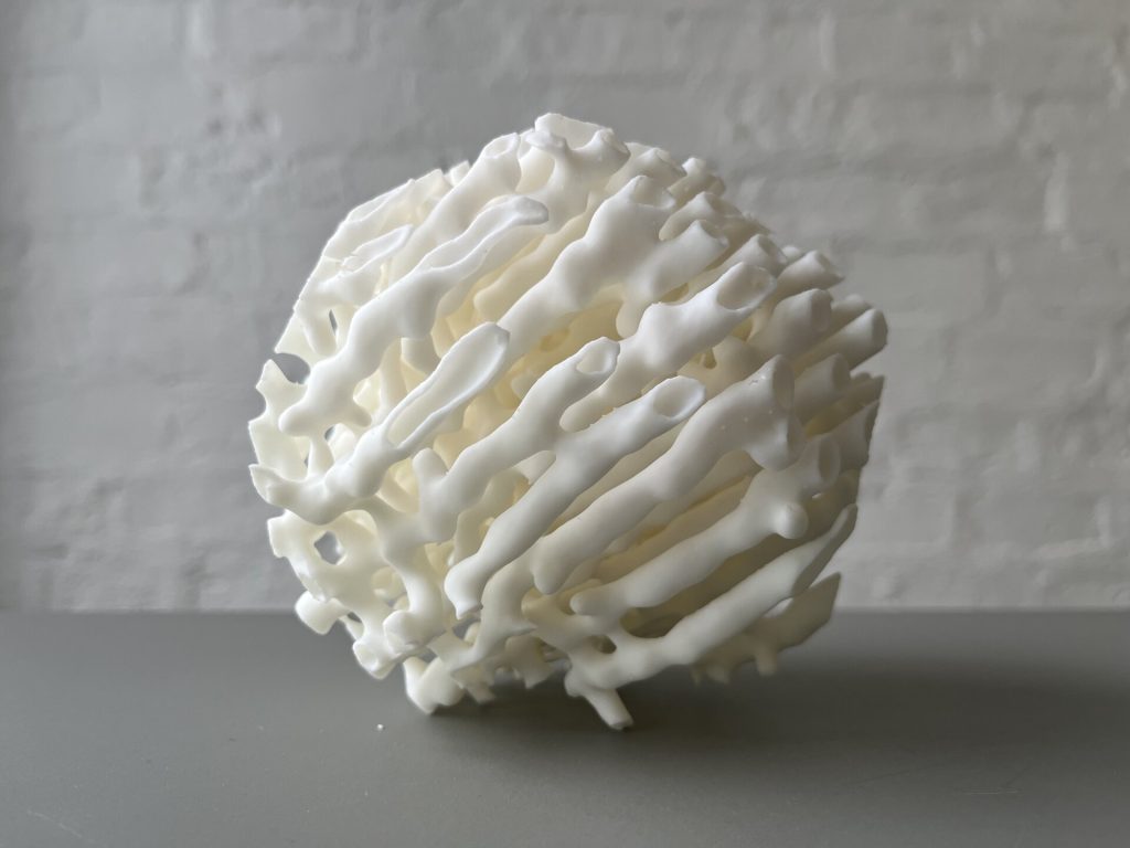 A white 3D-printed resin sphere with porous, coral-like structure sits on a gray surface against a white brick wall.
