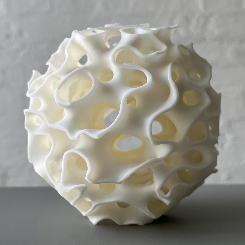 A white 3D-printed resin sphere with wavy, porous holes sits on a gray surface against a light wall.