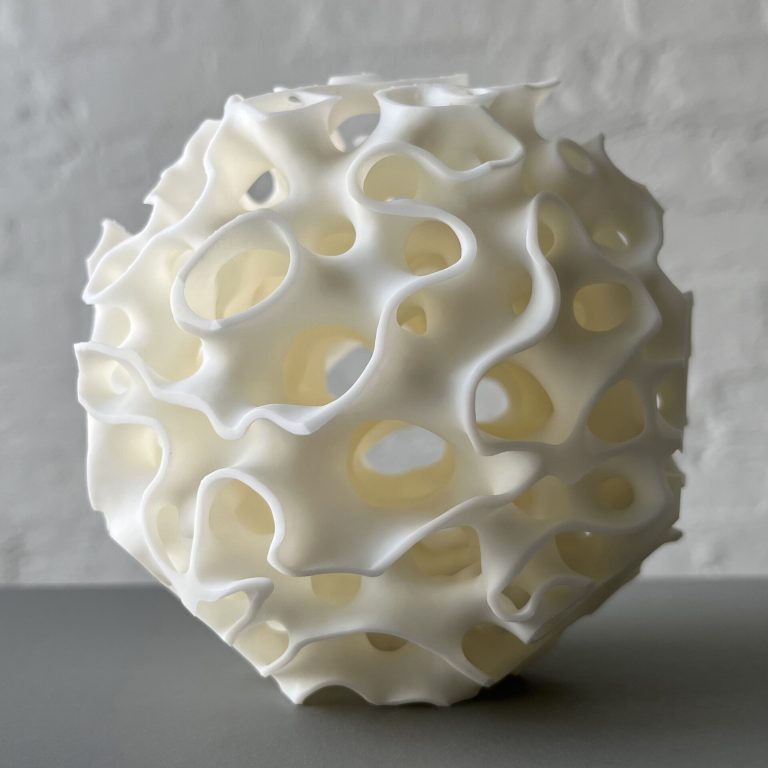 A white 3D-printed resin sphere with wavy, porous holes sits on a gray surface against a light wall.