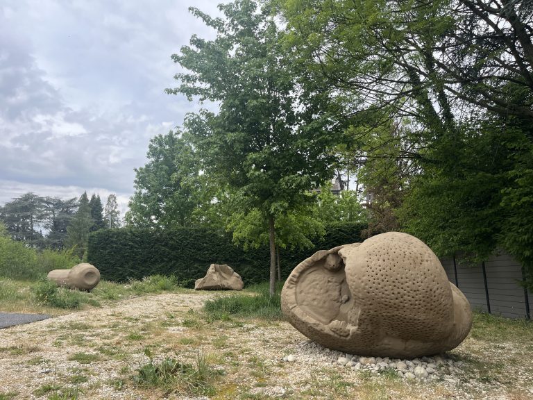 Three sand grain sculptures sit on a gravel path surrounded by trees and hedges.