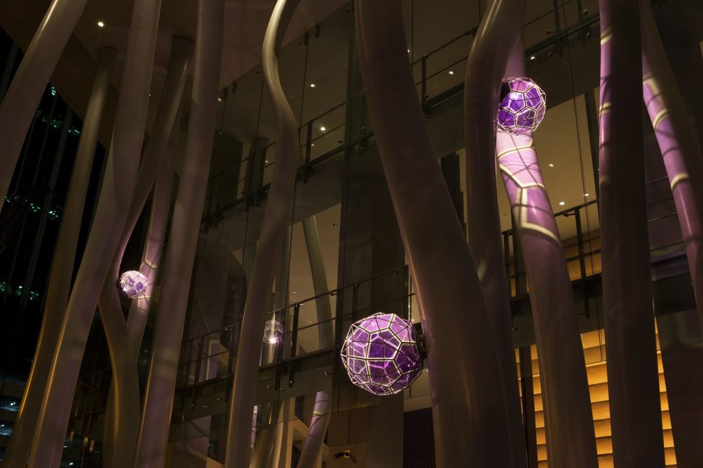 Suspended purple glass crystals hang between curved steel columns in a glass atrium.
