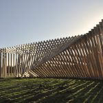 Wooden frames form a curved wall on grass with the sun behind them.
