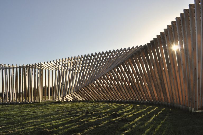 Wooden frames form a curved wall on grass with the sun behind them.