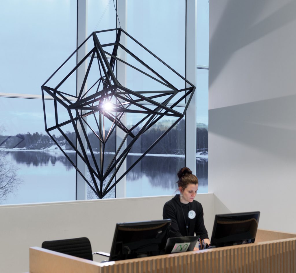 A light shines inside a suspended coated stainless steel geometric frame near a window.