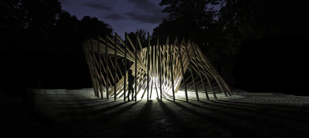A person stands inside a spiral of triangular wooden slats lit by sensor-controlled light at night.