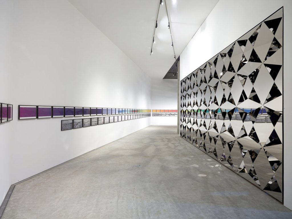 A stainless-steel mirror wall with diamond shapes stands in a gallery with framed art on the white walls.