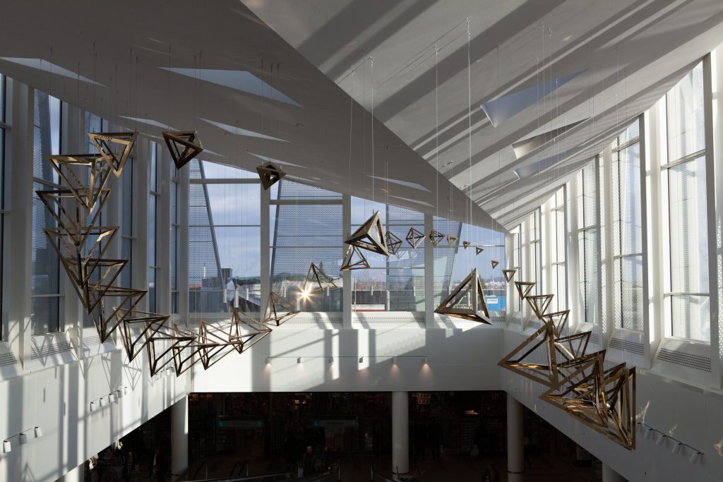Coated stainless steel tetrahedra hang from wires in a Möbius strip inside a sunlit atrium.