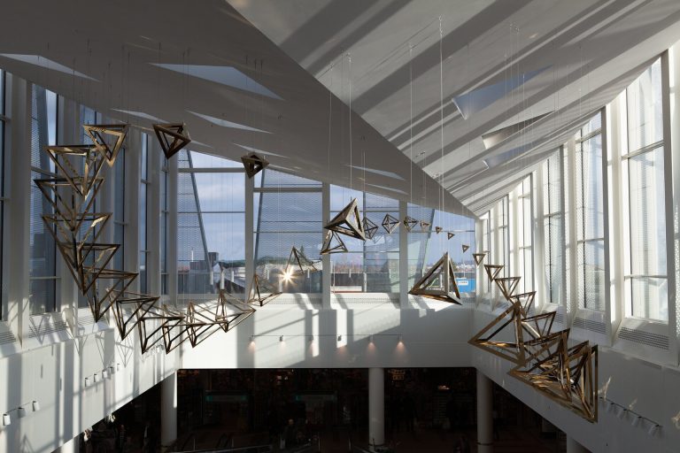 Coated stainless steel tetrahedra hang from wires in a Möbius strip inside a sunlit atrium.