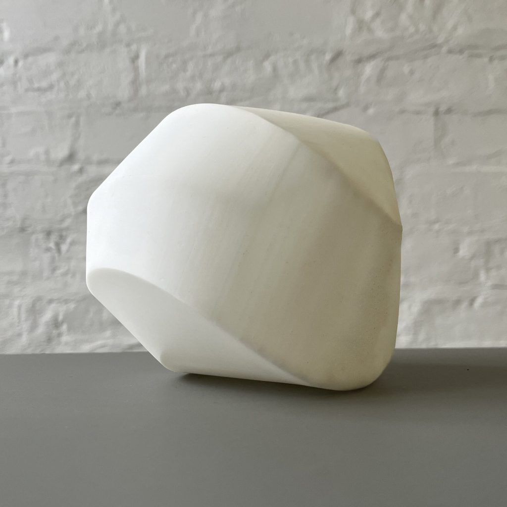 A white 3d printed resin polyhedron sits on a gray surface against a white brick wall.