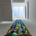 Colored glass triangles in stainless steel frames form a floor path in a white corridor.