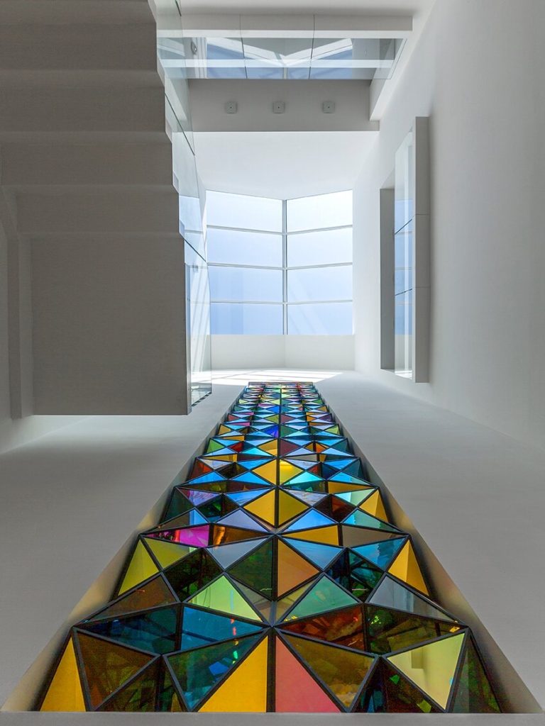 Colored glass triangles in stainless steel frames form a floor path in a white corridor.