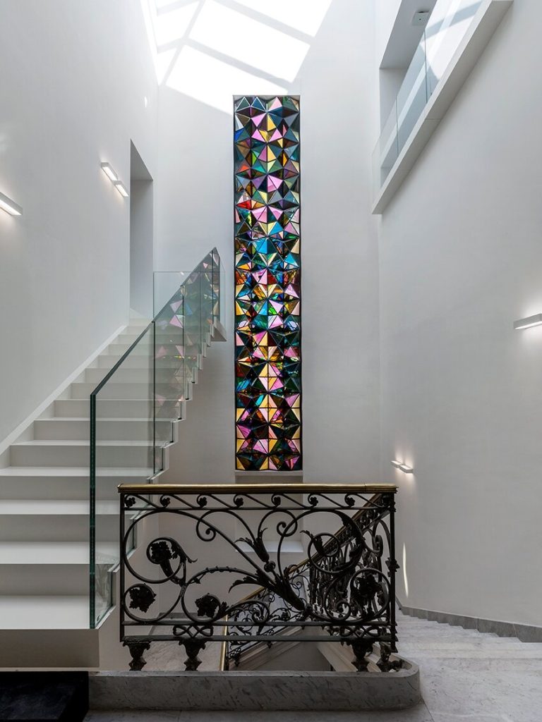 A vertical column of geometric window elements sits between a white staircase and a wrought-iron railing.