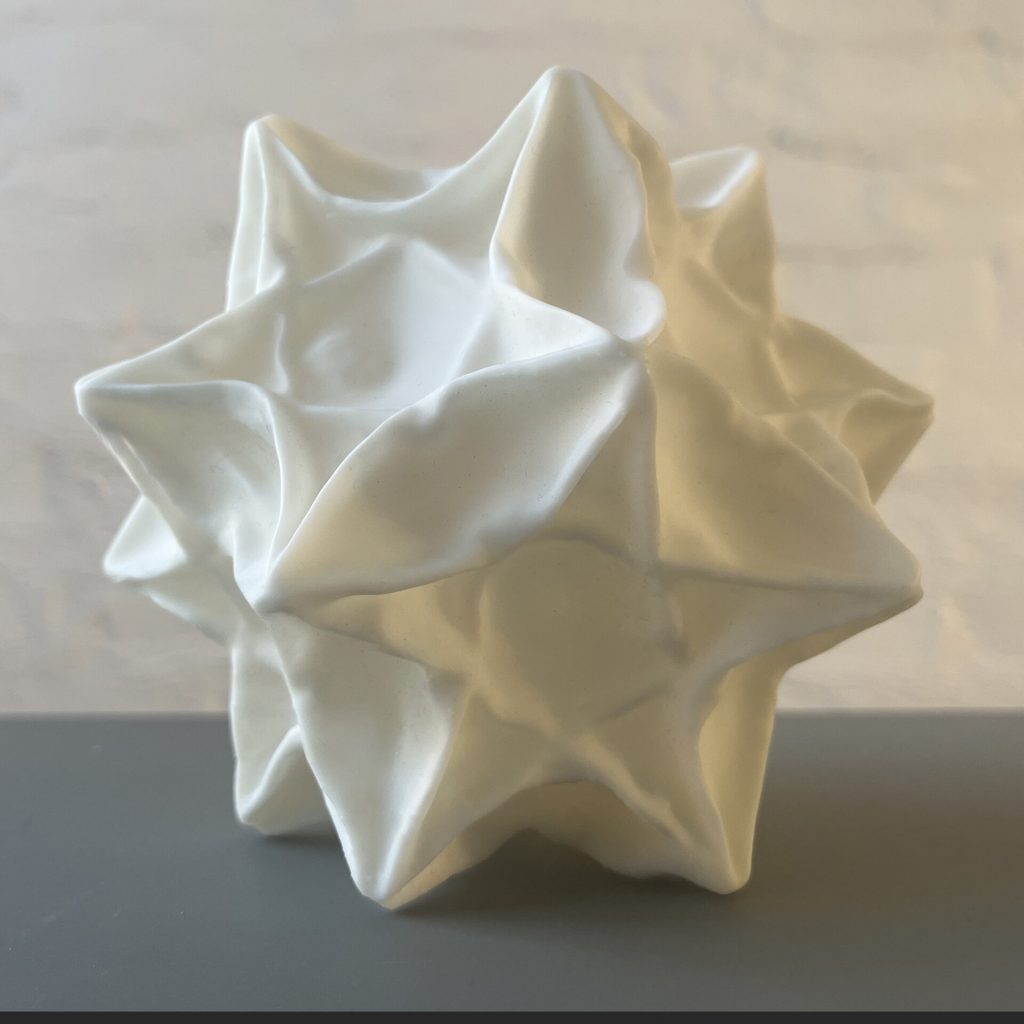 A white 3d printed resin stellated polyhedron sits on a gray surface.