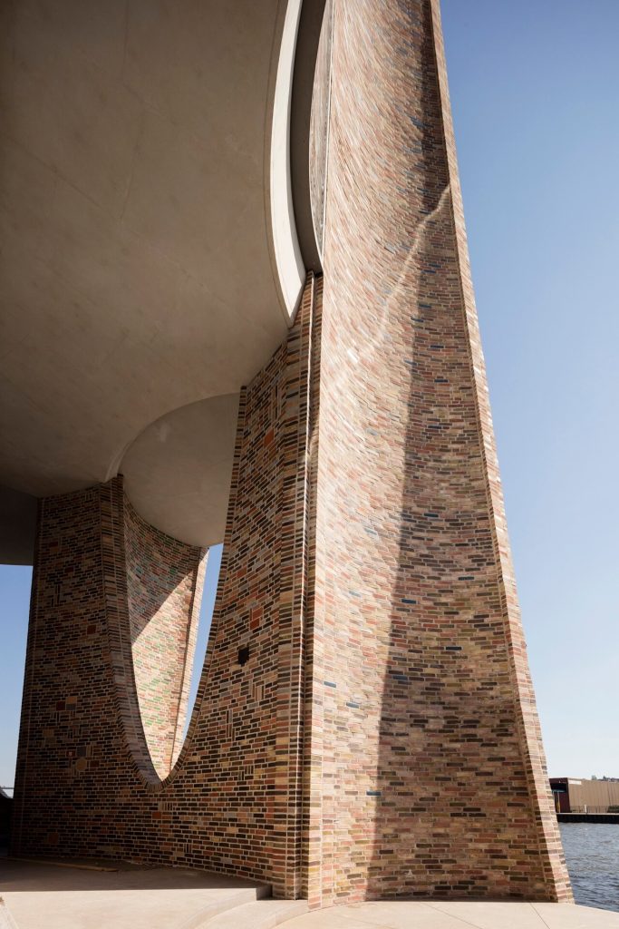 A curved brick facade with an ellipsoidal negative space sits under a concrete overhang.