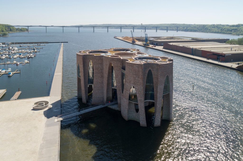 Brick cylinders with arched windows stand in the water, connected by a walkway.