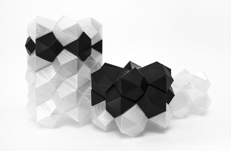 Black and clear 3D printed resin polyhedra interlock in geometric clusters on a white surface.