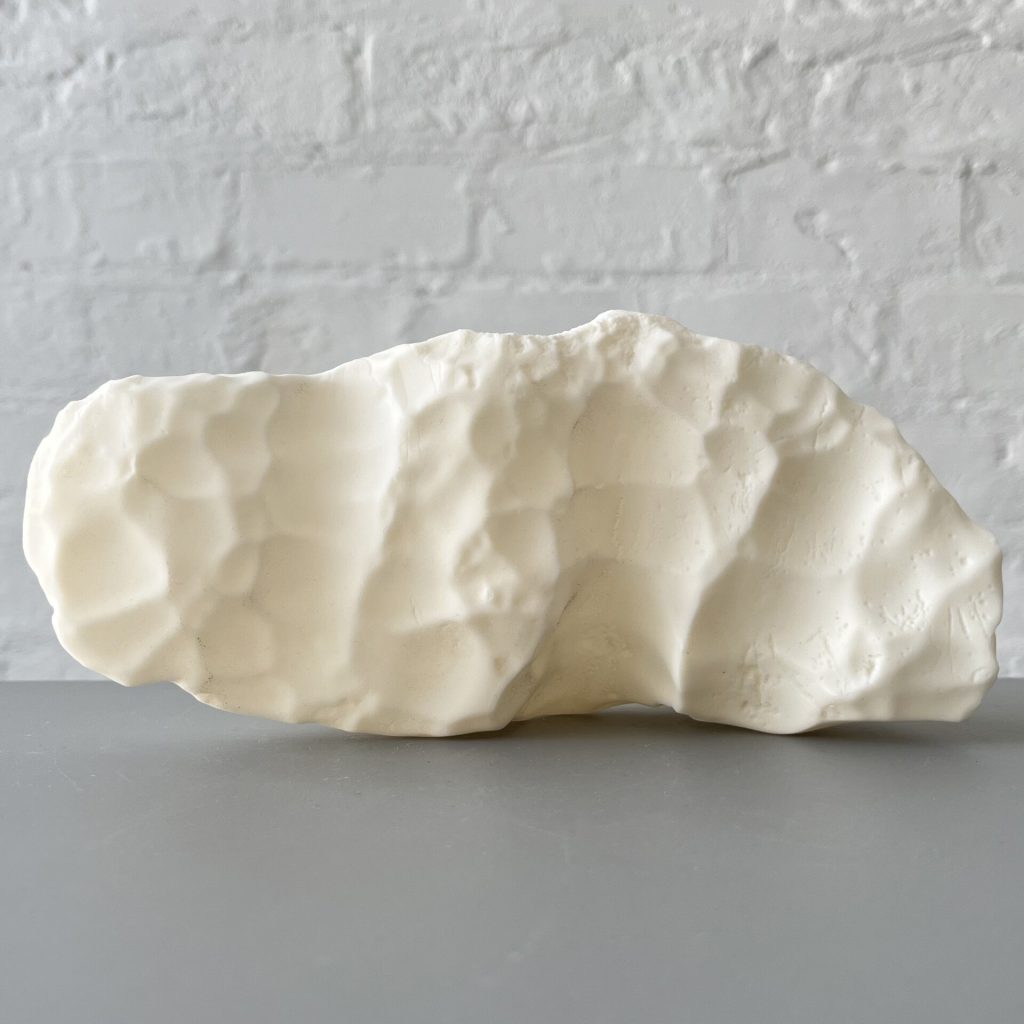 A white 3D printed resin object with a textured surface sits on a gray surface against a white brick wall.