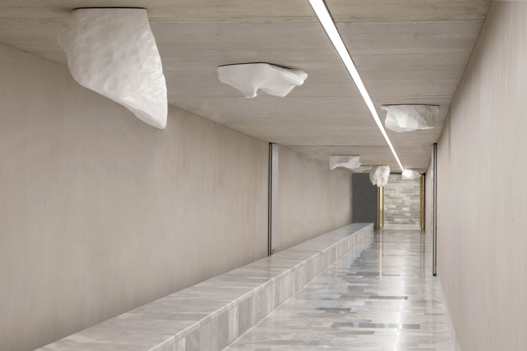 Seven Carrara marble blocks hang from the ceiling of a concrete tunnel with a marble bench.