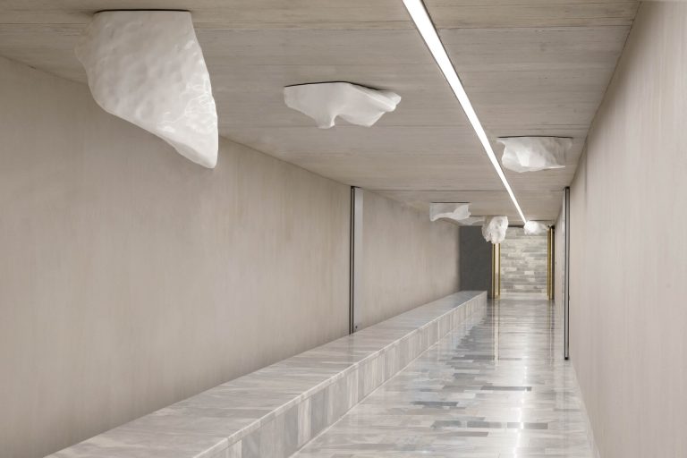 Seven Carrara marble blocks hang from the ceiling of a concrete tunnel with a marble bench.