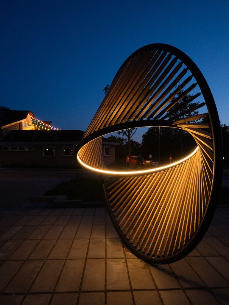 A painted stainless steel ring holds wooden components and an integrated lighting system on a paved surface.