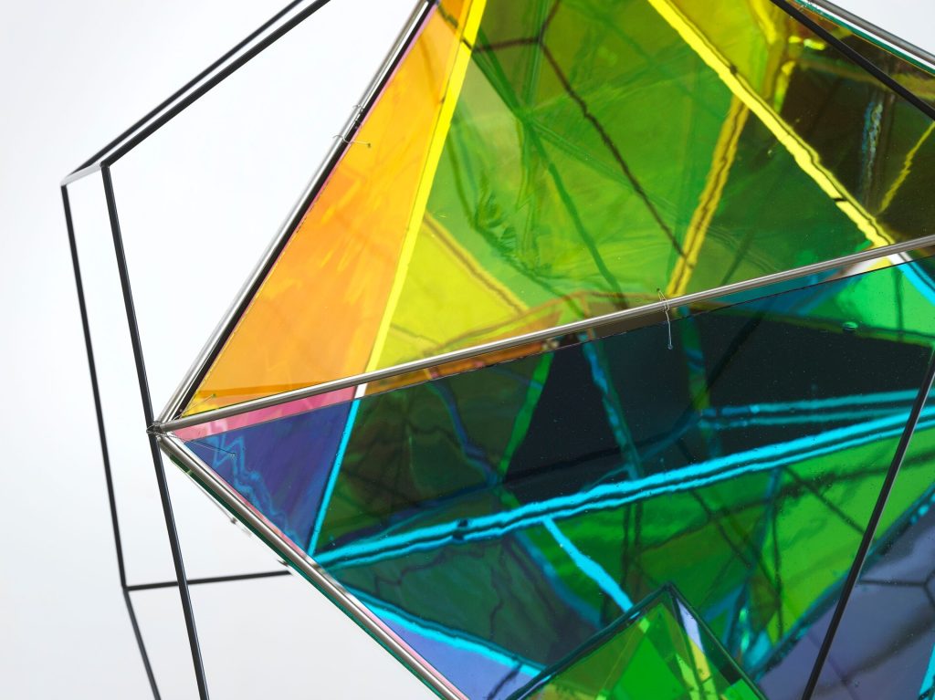 A black wire frame holds overlapping painted polyhedral elements in yellow, green, and blue.