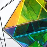 A black wire frame holds overlapping painted polyhedral elements in yellow, green, and blue.