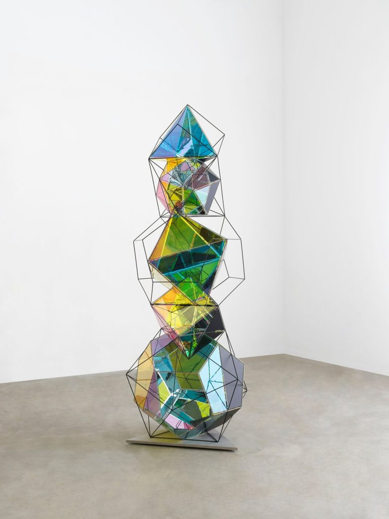 A stack of painted polyhedral elements sits on a metal base in a white room.