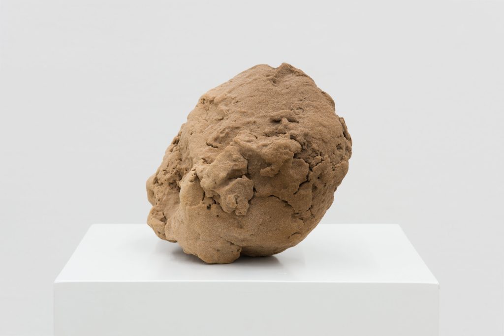 A 3D-printed sand sculpture with a rough, brown surface sits on a white plinth.