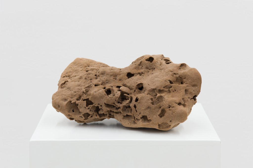A 3D-printed sand sculpture with porous texture sits on a white pedestal.