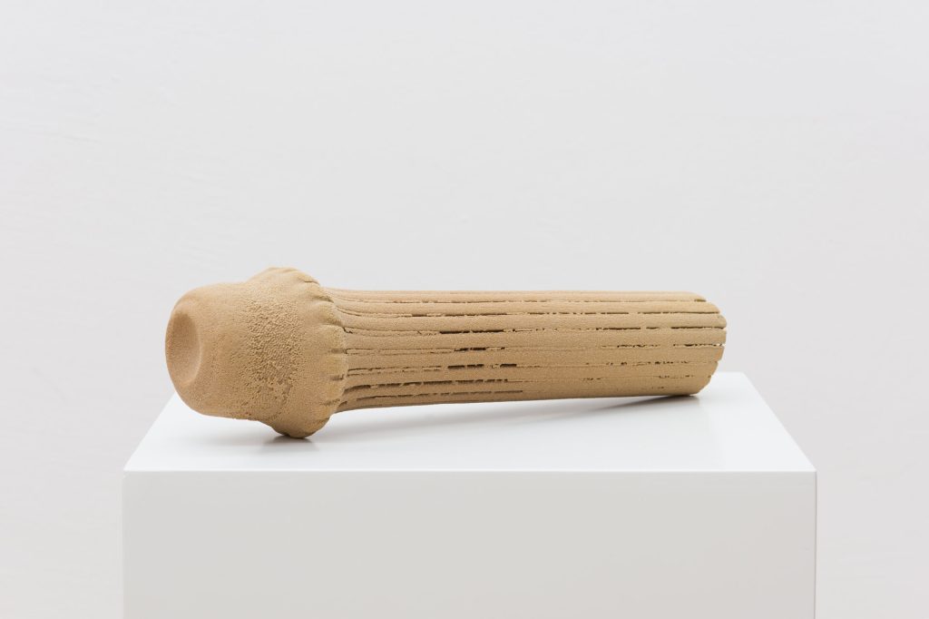 A 3D-printed sand sculpture with a rounded head and grooved shaft sits on a white plinth.