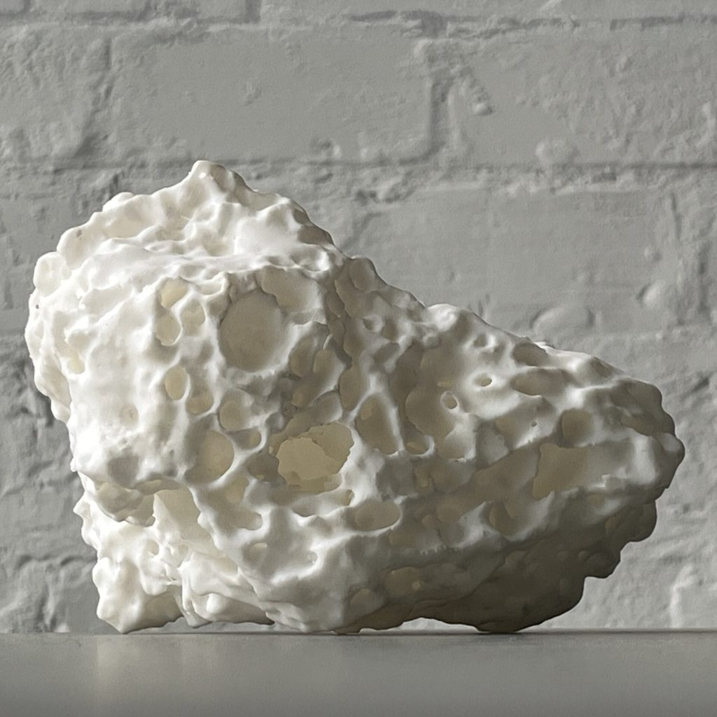 A white 3D printed resin object with porous holes sits on a surface against a brick wall.