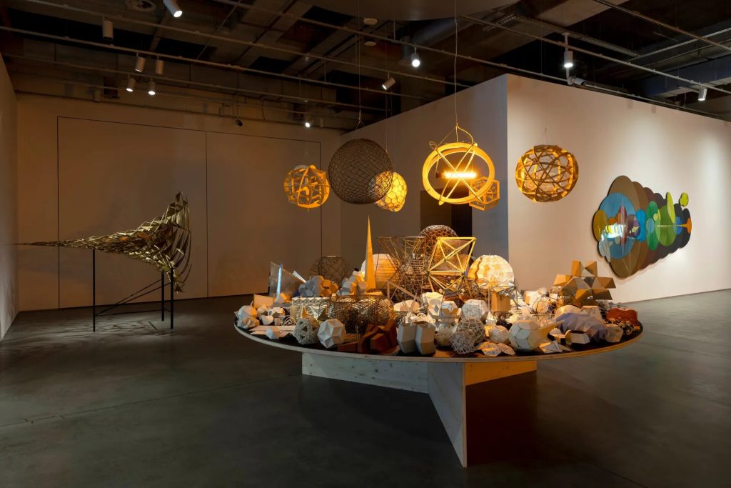 A circular wooden table holds geometric maquettes and prototypes while wireframe spheres hang above.
