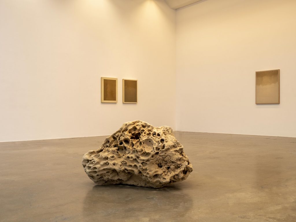 A porous 3D-printed sand sculpture sits on the floor before three framed sand paintings.