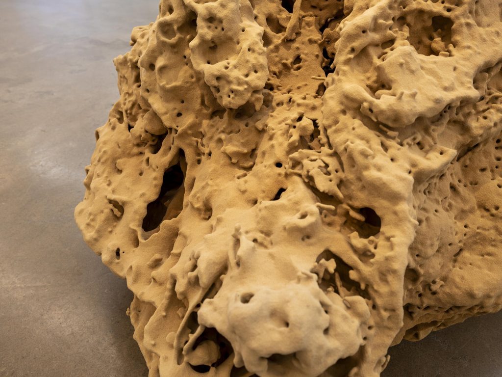 A 3D printed sand sculpture has a porous, tan surface with irregular holes and ridges.