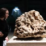 A porous 3d printed sand sculpture sits on a wooden pedestal while two people observe it.