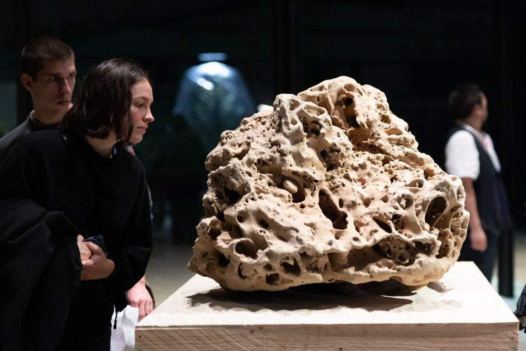 A porous 3d printed sand sculpture sits on a wooden pedestal while two people observe it.