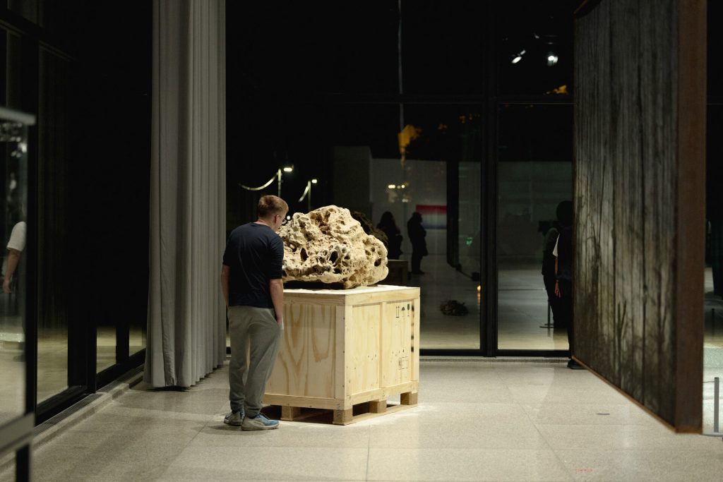A 3d printed sand sculpture sits on a wooden crate while a man looks at it.