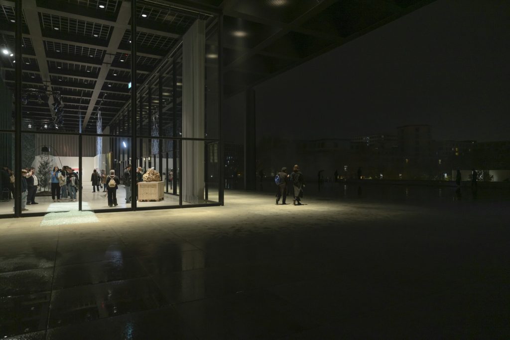 A 3d printed sand sculpture sits on a wooden crate inside a glass-walled gallery at night.