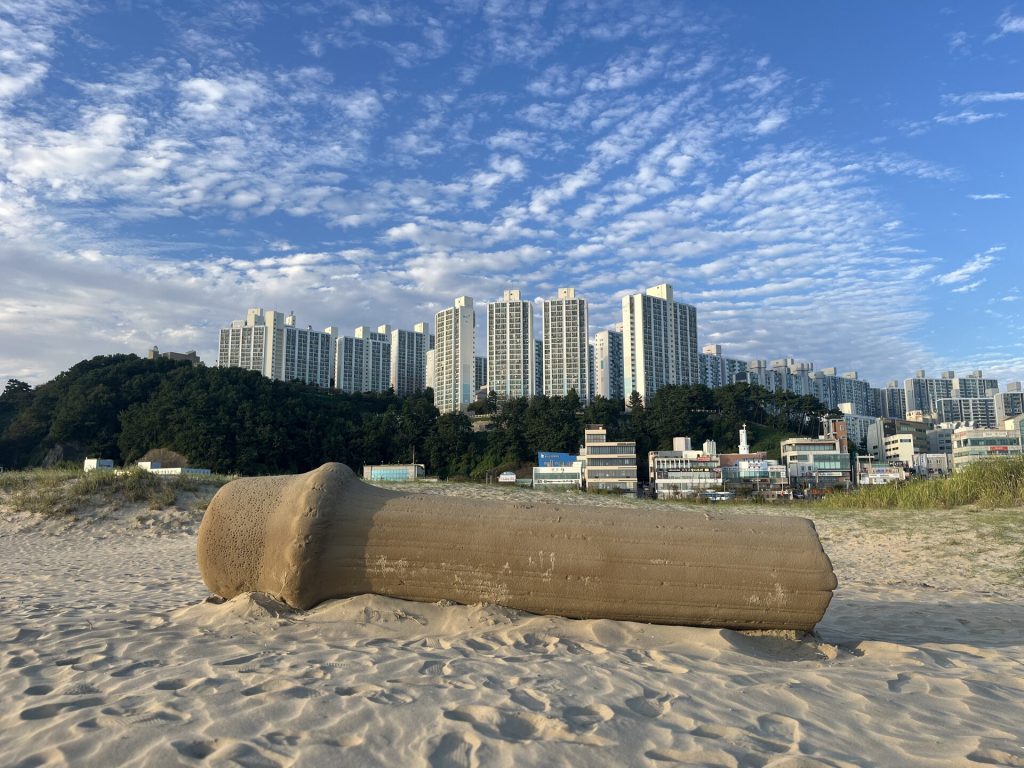 A sand sculpture rests on the beach with high-rise buildings in the background.