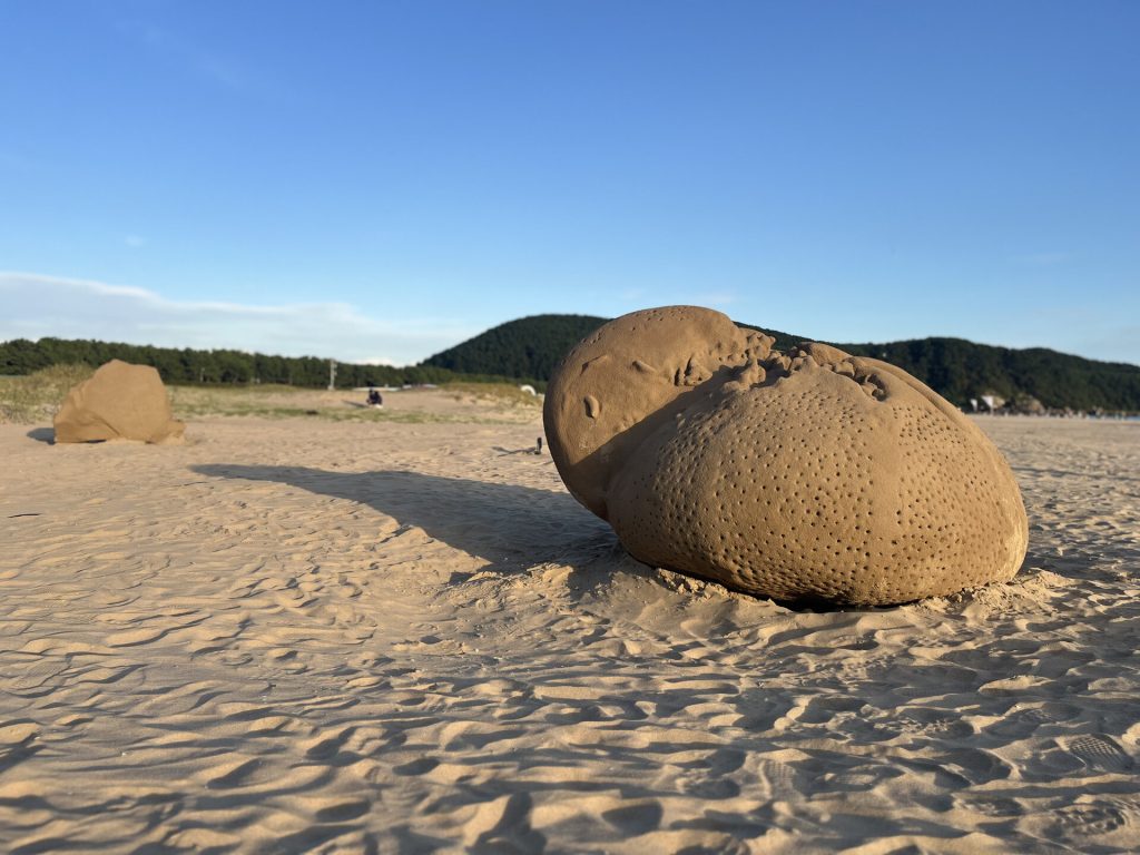 A large sand sculpture with pitted texture sits on a beach near a second sand form.