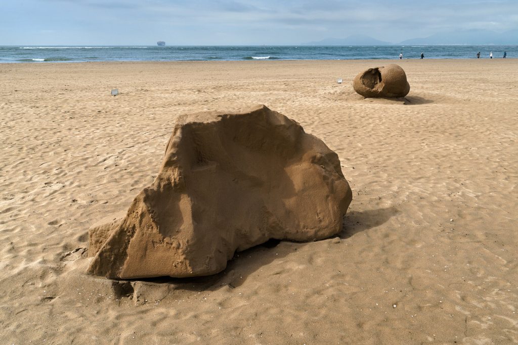 A large sand sculpture sits on a beach with a second sand form in the background near the ocean.