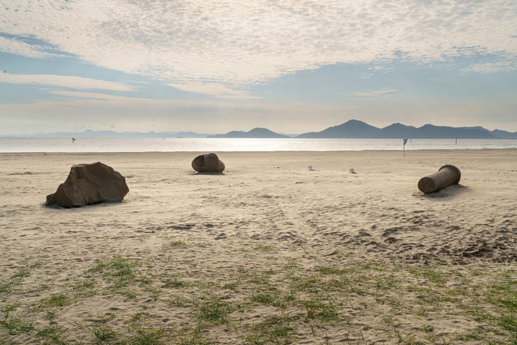 Three sand sculptures sit on a beach with mountains and water in the background.